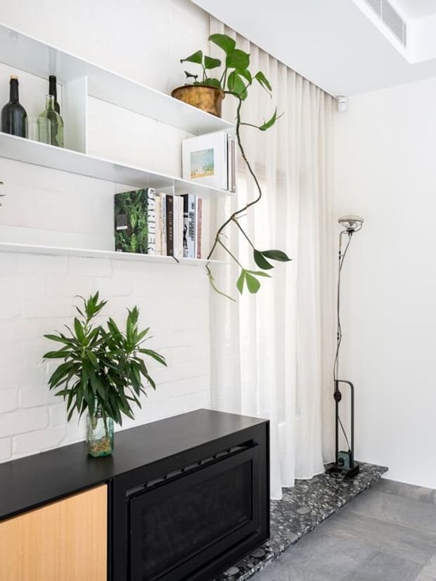Bright, refined interior corner featuring white painted brickwork, open white shelving with books and glassware, indoor plants, a black and timber built-in cabinet, sheer floor-to-ceiling curtains, and a dark patterned stone or terrazzo threshold beside a slim floor lamp.