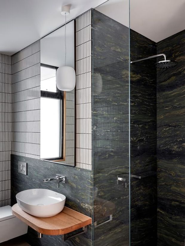 Contemporary bathroom featuring a wall-mounted white vessel basin on a rounded timber vanity, dark veined stone wall surfaces, a clear glass shower enclosure, white grid-pattern wall tiles, a black-framed window, and a sculptural pendant light.