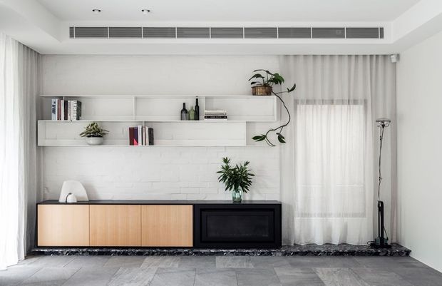 Bright contemporary living room featuring a long low built-in media console with light timber cabinetry and a black fireplace or media insert, set against a white painted brick feature wall. Floating white shelves display books, bottles and indoor plants, with sheer curtains, a tall floor lamp and grey stone-look flooring completing the calm minimalist scheme.