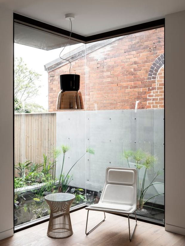 A compact, light-filled interior corner with floor-to-ceiling corner glazing overlooking a planted courtyard and an exposed brick wall. The space contains a modern pendant light, a sculptural lounge chair, and a small side table, with polished concrete screening panels and timber flooring creating a refined contemporary setting.