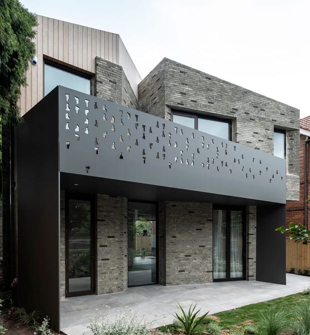 Street-facing view of a contemporary multi-level house with a dark perforated metal balcony enclosure, pale grey brick masonry walls, large dark-framed glazed doors and windows, and vertical light-coloured cladding to the upper facade. A paved terrace and landscaped garden sit in front of the building.