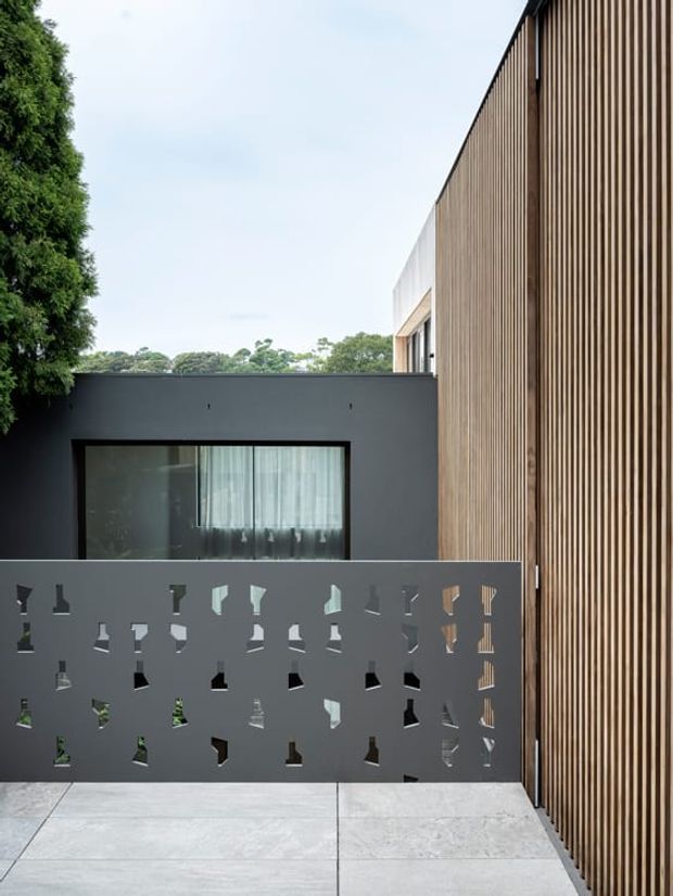 Contemporary residential exterior courtyard view featuring a tall vertical timber screen facade beside a dark rendered wall with a large window. A perforated dark privacy wall and pale paved terrace occupy the foreground, with greenery visible beyond.
