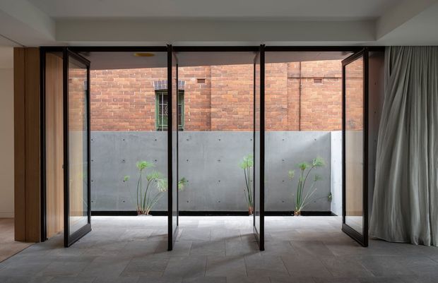 A refined interior space opening onto a small courtyard through full-height black-framed glazed folding or pivot doors. The room has a smooth light concrete ceiling, grey large-format tiled flooring, warm timber wall panels, and full-height grey curtains. Outside, a concrete planter wall is backed by exposed red brickwork with sparse greenery, creating a calm indoor-outdoor connection.