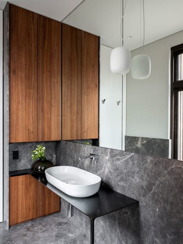 A contemporary bathroom featuring a floating white vessel basin on a dark benchtop, walnut-toned vertical cabinetry, grey marble-look wall and floor finishes, a large mirror, black-framed window, and suspended rounded pendant lights.