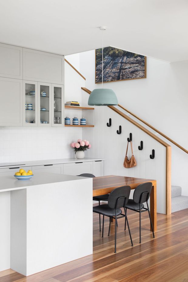 Bright, compact open-plan kitchen and dining area beneath a staircase. The space features white built-in cabinetry with glass-front display doors, a pale benchtop and island, white tiled splashback, warm timber flooring and dining table, black dining chairs and wall hooks, and a muted sage pendant light. A timber handrail and stair balustrade frame the background, with framed artwork above.