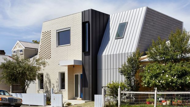 Street-facing exterior view of a contemporary multi-level residence with a strongly geometric composition. The home combines pale textured masonry, dark vertical cladding, and a steep folded light-metal roof or facade plane with inset windows. A small front entry, low fencing, garden planting, and neighbouring residential buildings are visible.