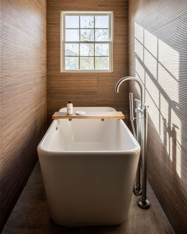 A narrow, warmly lit bathroom centered on a freestanding white bathtub beneath a divided-pane window. The walls are clad in fine horizontal brown textured tiles or ribbed panels, with dark stone-look floor tiles, a timber bath ledge, and an exposed chrome freestanding bath filler and hand shower. Strong sunlight casts linear window shadows across the right wall.