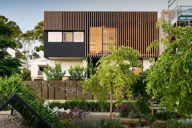Contemporary multi-storey residential facade viewed from a landscaped front garden. The upper level features a strong vertical timber-look screening system, dark cladding, and recessed glazing, while the lower level is light-coloured with a partially visible boundary fence. Dense ornamental planting and a pedestrian path frame the building.