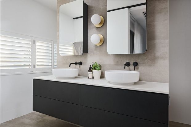 Contemporary double-vanity bathroom featuring two white vessel basins on a long white benchtop, dark matte floating cabinetry, concrete-look wall and floor finishes, rounded rectangular mirrors, globe wall sconces, and white plantation shutters.
