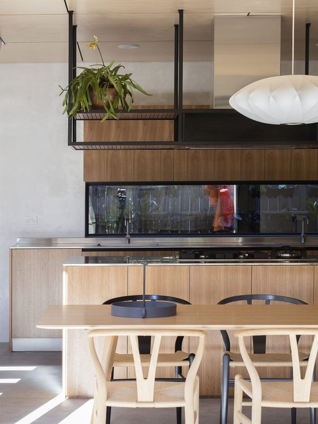 A refined contemporary kitchen and dining area with extensive warm timber joinery, a long island or dining counter, black metal-framed overhead shelving, stainless-steel work surfaces, and a dark glass splashback. Sculptural white pendant lighting, timber dining chairs, and a hanging plant add visual interest. The space is bright and understated, with a blend of natural wood and black detailing.
