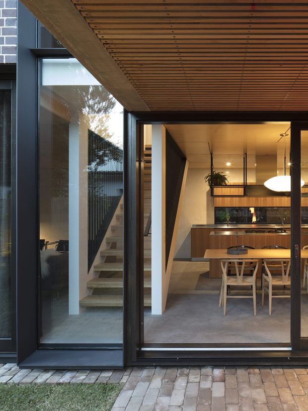 View from an outdoor paved area through large dark-framed glazing into a contemporary open-plan dining and kitchen space. The interior features a timber dining table and chairs, dark timber kitchen cabinetry, a long island or counter, pendant lighting, concrete flooring, and a staircase visible to the left. A slatted timber soffit extends over the glazed opening, with exposed brickwork and planted greenery adding texture.