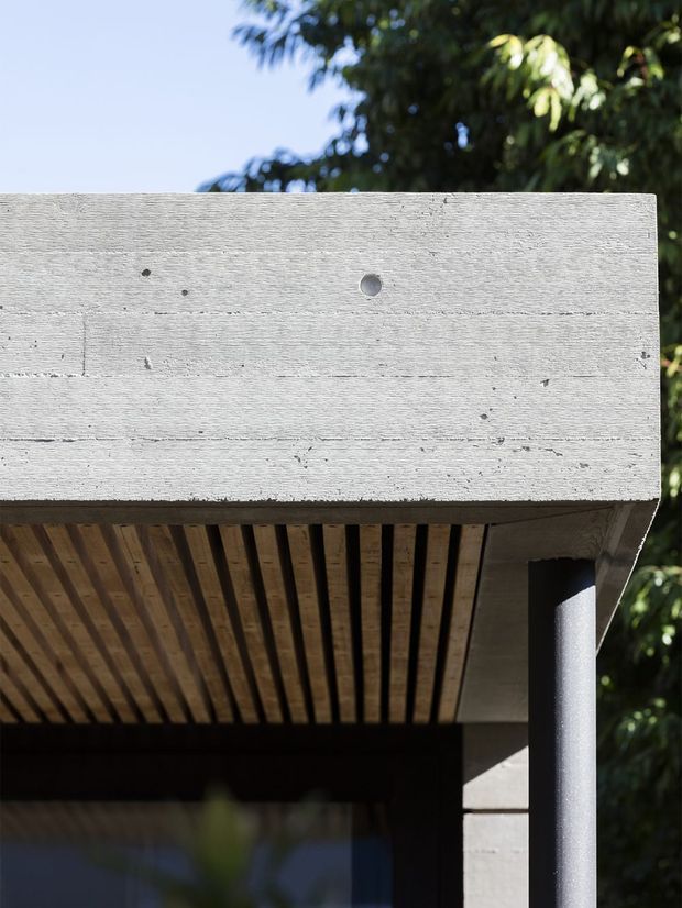 Close-up exterior architectural detail showing a flat concrete roof slab with a pale textured finish, supported by a dark cylindrical column. The underside features closely spaced timber slats forming a soffit, with dark glazing or an entry opening behind. Vegetation and sky are visible in the background.