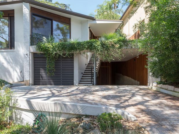 A contemporary multi-level residential facade viewed from the driveway, featuring white textured rendered walls, dark-framed glazing, timber detailing, a flat roof, an integrated garage, and a covered carport or entry beneath a planted balcony. An external staircase rises between the garage and upper-level entry, with mature landscaping surrounding the concrete driveway and terrace.