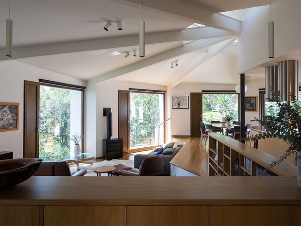 A bright, open-plan residential interior viewed from behind a timber kitchen or cabinetry counter. The space combines a living area and dining area, with large timber-framed windows, exposed sloping ceiling forms, multiple skylights, and a contemporary timber kitchen island or bench. Neutral white walls and ceilings contrast with warm timber joinery and dark furnishings. Indoor plants and views to greenery add a natural feel.