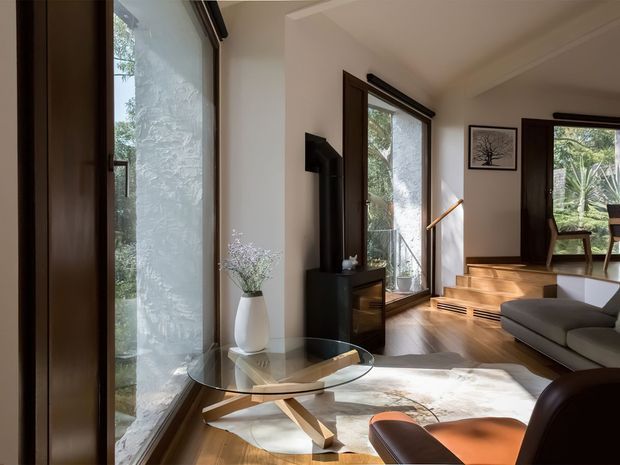 A sunlit contemporary living room with extensive dark-framed glazing, warm timber flooring, and a raised adjoining area. The room contains a sculptural glass coffee table, neutral sectional sofa, leather lounge chair, dark media console, and a pale rug. Abundant garden views and natural light create an airy, refined atmosphere.
