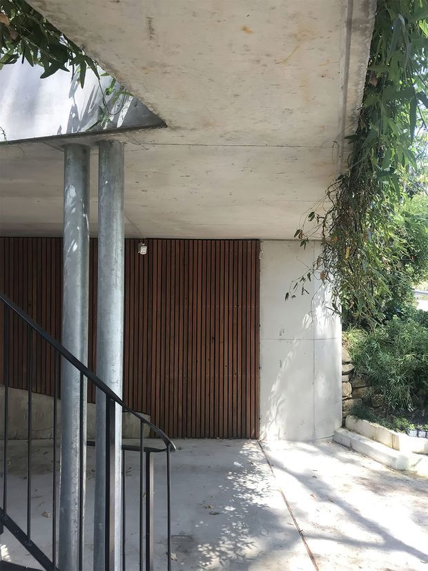 A shaded modern building entrance or covered outdoor passage with raw concrete slab roofing, cylindrical concrete columns, a black metal handrail, and a large vertical timber slatted door or screen. Dense climbing vegetation and a small landscaped retaining bed border the right side, creating a strong indoor-outdoor character.
