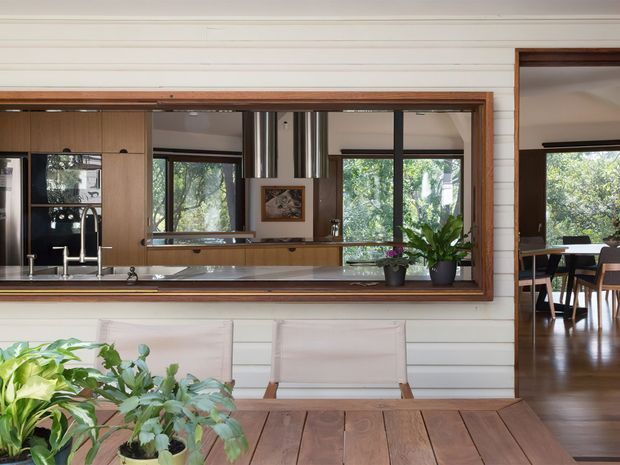 View through a large timber-framed interior pass-through window into a warm, open-plan kitchen and dining area. The kitchen features dark cabinetry, stainless-steel appliances and rangehood, a light benchtop, and extensive glazing overlooking greenery. White horizontal wall panelling surrounds the opening, while a timber outdoor or indoor table and potted plants occupy the foreground.