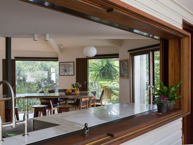 A bright open-plan kitchen and dining area viewed through a large interior pass-through or servery opening. The kitchen features a pale stone island benchtop, stainless-steel sink and tapware, white cabinetry, and substantial dark timber-framed joinery. Beyond it, a timber dining table and chairs sit beside expansive windows and glazed doors overlooking lush greenery. The room has a warm, refined, indoor-outdoor character with abundant natural light.