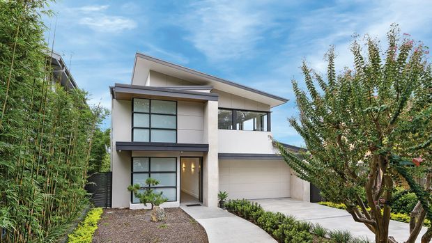 Exterior view of a contemporary two-storey residence with a simple low-pitched roof, white rendered walls, dark metal-framed glazing, a projecting upper-level balcony or canopy, and an integrated double garage. A paved path leads to the recessed entry, with landscaped planting around the frontage.