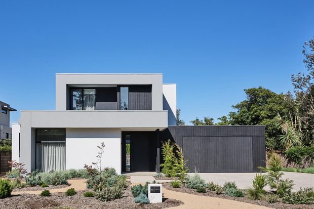 Street-facing view of a contemporary two-storey residence with a crisp white rendered facade, dark vertical garage cladding, deep charcoal window and door frames, and a flat roofline. The home is set behind a low-maintenance landscaped garden with gravel, clipped shrubs, and native-style planting.