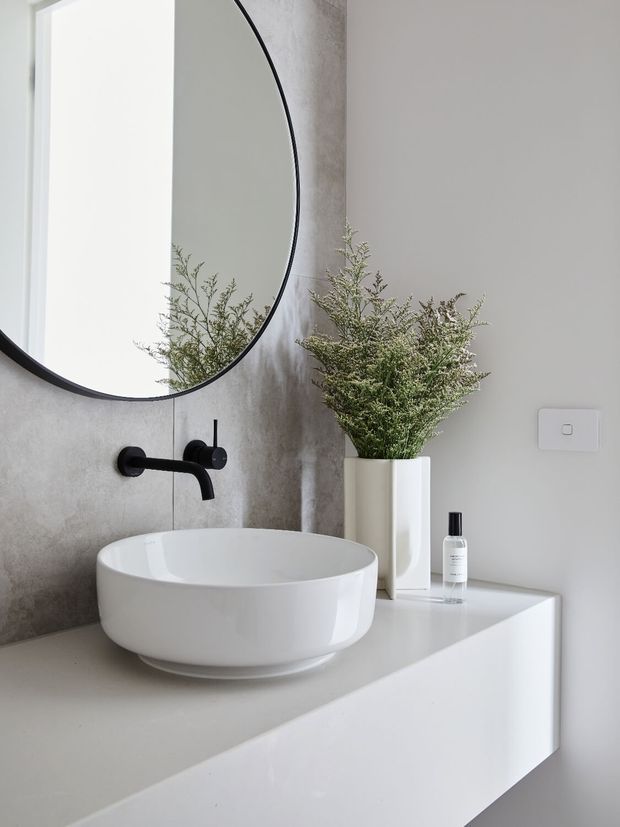 A minimalist bathroom vanity featuring a white vessel basin on a crisp white floating cabinet, a matte black wall-mounted tap, a large round black-framed mirror, and a tall white vase with greenery. The wall has a subtle grey textured finish, creating a clean, refined contemporary setting.