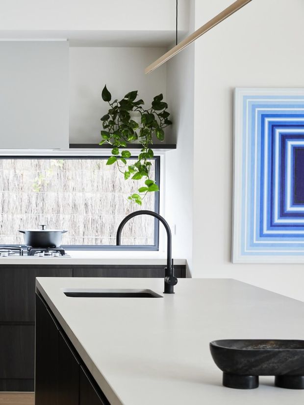 Bright contemporary kitchen interior with a large white island and integrated black sink, matte black gooseneck tap, dark cabinetry, a rear cooking zone, and a window above the benchtop. A trailing indoor plant hangs from the upper cabinetry, while blue geometric artwork adds a strong accent to the white walls. A slim linear pendant light runs overhead.