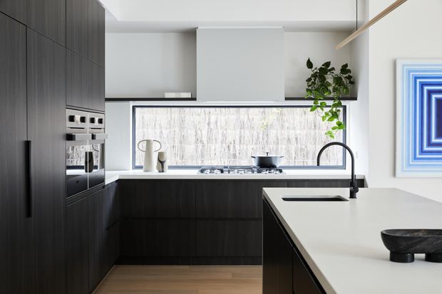 A refined contemporary kitchen with dark timber cabinetry, integrated stainless-steel appliances, a pale stone island benchtop, and a large textured stone or marble-look splashback beneath a wide window. The island includes an inset sink and sculptural dark decorative objects, while a potted plant and framed blue artwork add subtle accents.