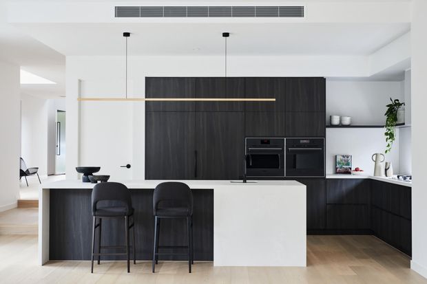A bright, contemporary open-plan kitchen with a large white island and two dark upholstered bar stools. Full-height dark timber-look cabinetry houses integrated ovens, while matching lower cabinetry extends along the rear wall beneath a white benchtop. Minimal linear pendant lighting, pale timber flooring, white walls, and concealed ceiling ventilation create a clean, refined interior.