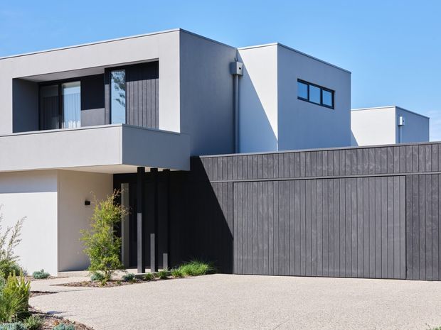 A contemporary multi-level residential facade with clean geometric volumes, pale rendered walls, dark vertical timber-look cladding, a large matching garage door, recessed entry, and projecting upper-level balcony. The home is set behind a light gravel driveway with sparse low landscaping under a clear blue sky.