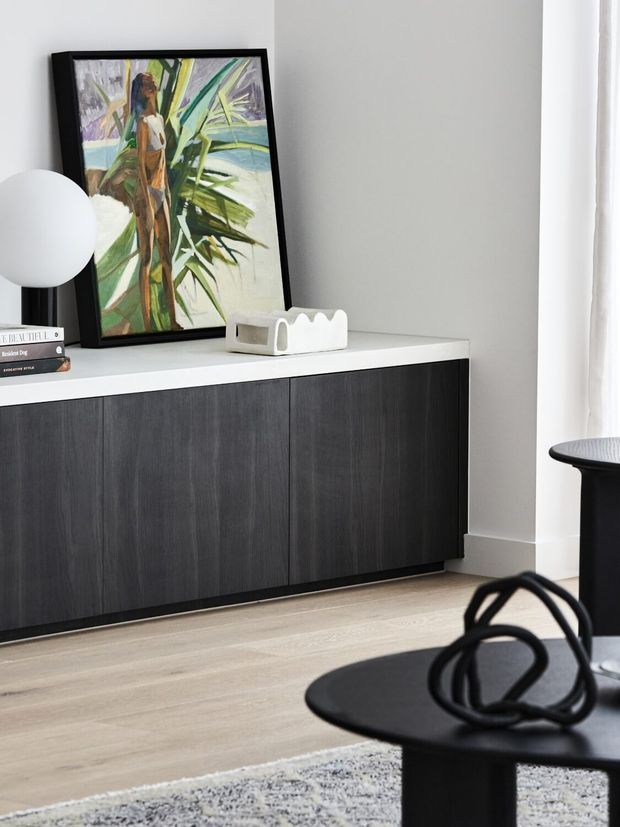 A refined contemporary living room vignette featuring a long dark timber-finish sideboard with a white countertop, pale timber flooring, white walls and curtains, and minimalist black furniture. A framed botanical artwork, sculptural white ornament, spherical lamp, and black coffee table add restrained decorative accents.
