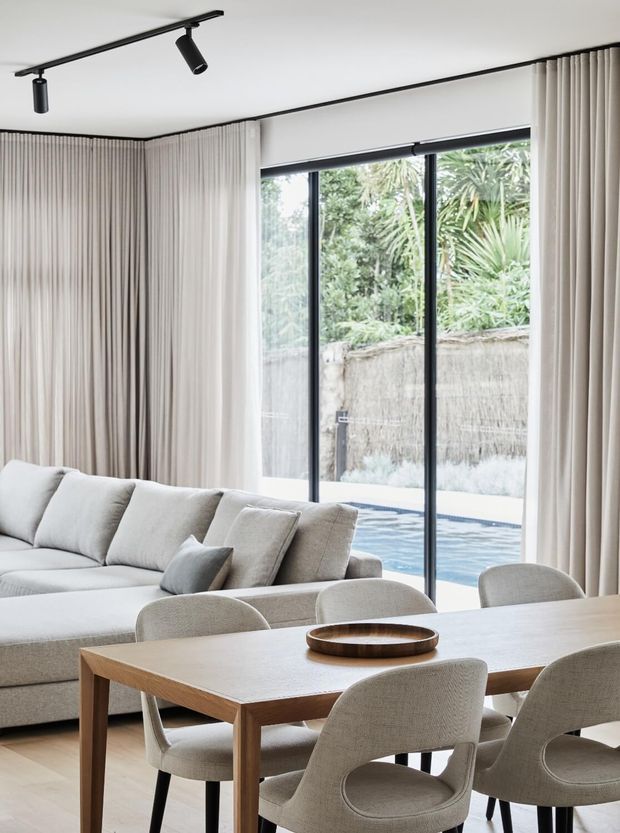 A bright, refined open-plan living and dining area with a neutral palette. A large light-toned sectional sofa sits beside a timber dining table with upholstered chairs. Full-height curtains frame expansive black-framed glazing, overlooking a small swimming pool and lush garden. Track lighting runs across the smooth white ceiling.