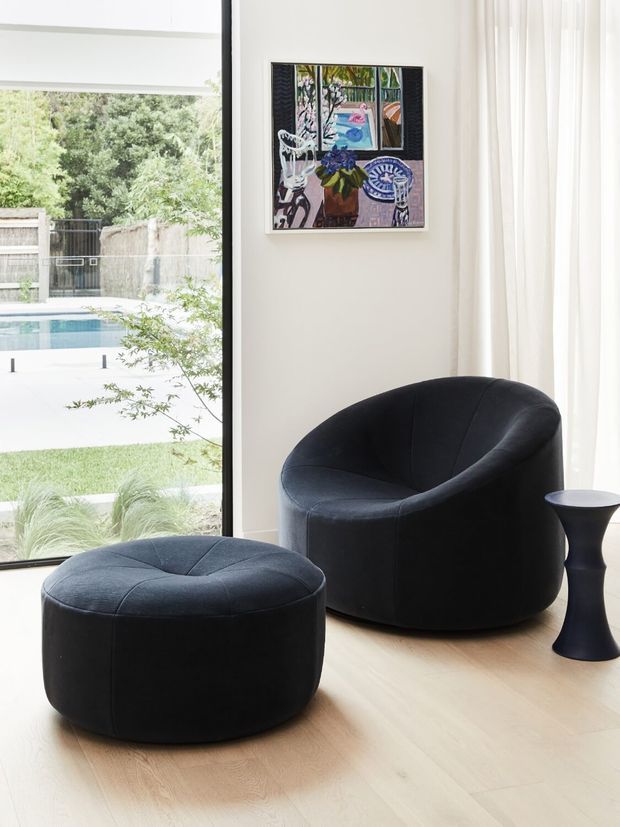 Bright, refined sitting area with a sculptural black upholstered lounge chair and matching round ottoman on a pale timber floor. Full-height glazing opens to a landscaped garden and swimming pool, while sheer curtains and colourful framed artwork add softness and visual interest. A small black pedestal side table completes the composition.