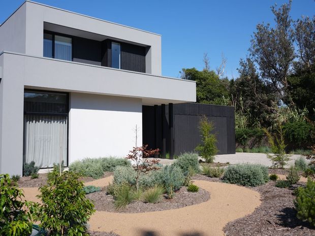 Exterior view of a contemporary two-storey flat-roof home with crisp white rendered walls, dark framed glazing, a projecting upper-floor volume, and a black vertical-slat privacy gate or screen. The front garden uses a low-maintenance drought-tolerant landscape with gravel or mulch beds, pale decomposed-granite pathways, ornamental grasses, shrubs, and young trees.