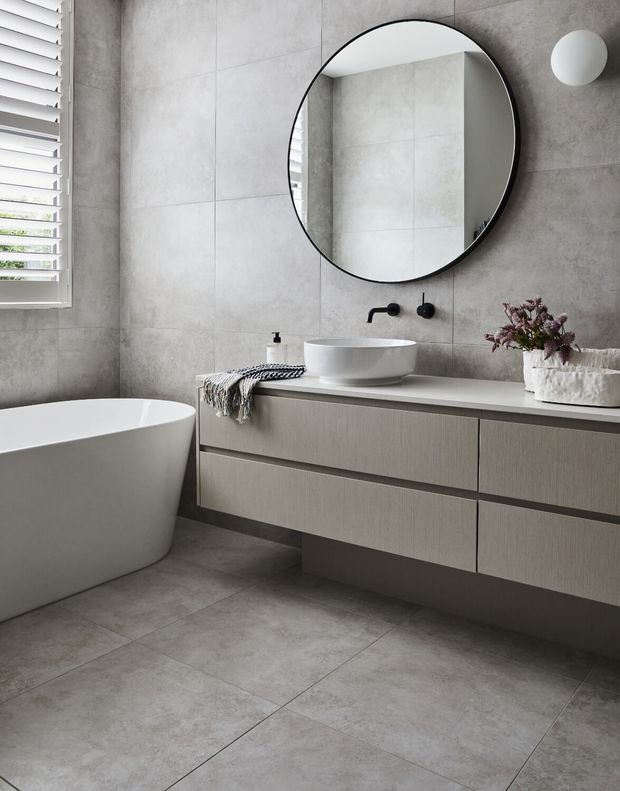Contemporary bathroom featuring large-format grey concrete-look wall and floor tiles, a floating light wood vanity with integrated drawers, a white vessel basin, matte black wall-mounted tapware, a freestanding white bathtub, round black-framed mirror, and white horizontal window blinds. The space has a calm, refined and minimalist aesthetic.