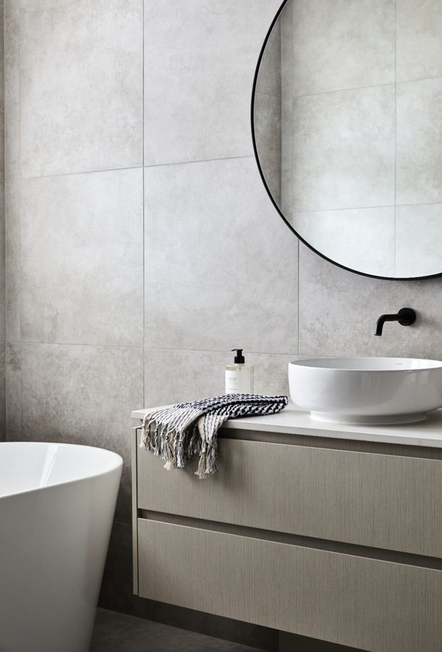 A contemporary bathroom with large light-grey concrete-look wall tiles, a freestanding white bathtub, wall-mounted vanity with textured grey cabinetry, white vessel basin, matte black wall-mounted tapware, and a large round mirror. A monochrome woven textile adds subtle texture.