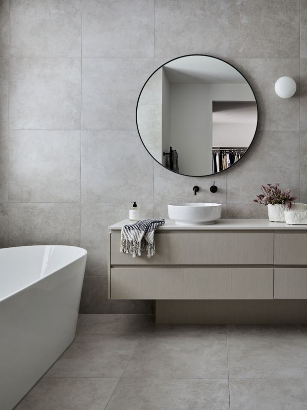 Contemporary bathroom with large grey concrete-look wall and floor tiles, a freestanding white bathtub, floating light timber-toned vanity, white vessel basin, round black-framed mirror, and minimalist black wall-mounted fixtures. The mirror reflects open shelving and hanging clothing from an adjacent space.