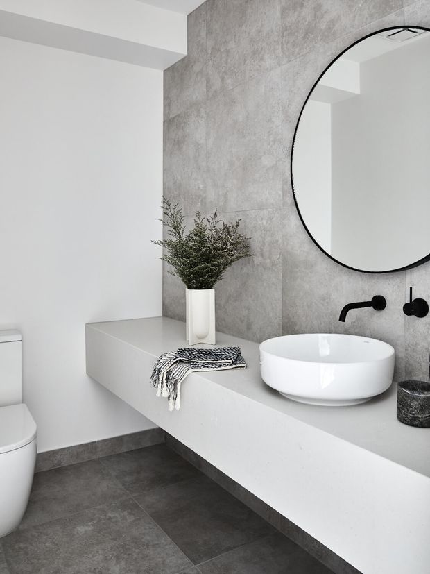 A minimalist contemporary bathroom featuring a floating white vanity with an integrated counter, round white vessel basin, matte black wall-mounted tapware, and a large round black-framed mirror. The walls and floor use grey concrete-look finishes, creating a clean monochromatic scheme with a small amount of greenery and soft textile decor.
