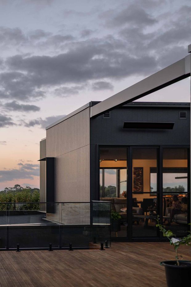 Exterior architectural photograph at dusk showing a contemporary multi-level building with dark rendered walls, pale vertical cladding, strong angular roofline, and large black-framed glazed doors opening onto a broad timber deck. A glass balustrade borders the elevated outdoor area, with warm interior lighting visible through the glazing and a cloudy evening sky beyond.