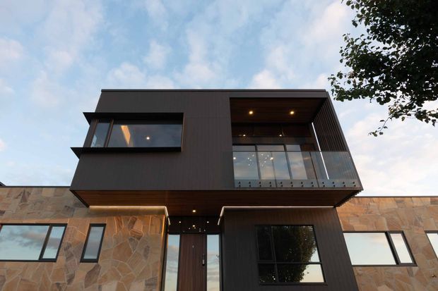 Low-angle exterior view of a contemporary multi-level residence with projecting upper volumes, dark vertical cladding, extensive glazing, recessed balcony or terrace, and stone-clad lower walls. The facade combines strong geometric forms with warm timber-toned soffits and integrated lighting.