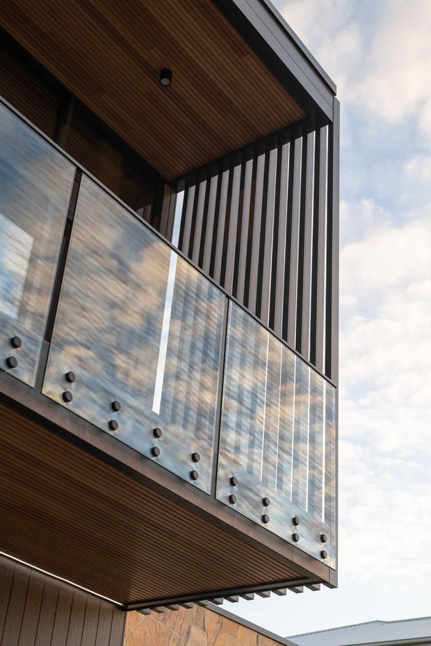 Close-up upward view of a contemporary building exterior featuring a projecting balcony with clear glass balustrades, dark metal framing, vertical dark-and-light facade fins, and warm timber-lined soffits. Stone wall cladding and a roof edge are partially visible below, with sky in the background.