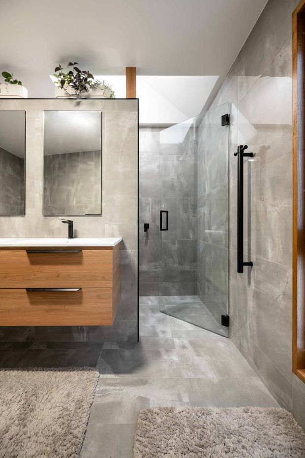A contemporary bathroom with a walk-in glass shower, grey stone-look tiled walls and floor, a floating timber vanity with a white benchtop, black tapware and shower hardware, frameless mirrors, and a skylight bringing in natural light. Small rugs and indoor plants add softness and warmth.