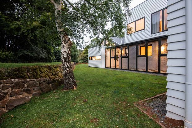 Exterior view of a contemporary multi-level residential building set within a landscaped garden. The facade combines horizontal white weatherboard cladding, dark brickwork, extensive black-framed glazing, and a projecting upper storey. A moss-covered stone retaining wall and mature birch tree frame the lawn, while warm interior lighting glows through the windows.
