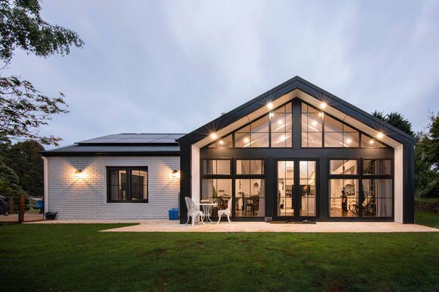 Exterior view of a single-storey house at dusk, featuring a dark-framed glazed gable extension with extensive floor-to-ceiling windows and warm interior lighting. The original light brick-clad building sits to the left, with a low-pitched roof incorporating solar panels. A lawn and trees surround the property.
