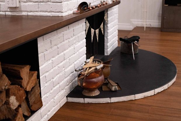 Close-up interior view of a traditional white-painted brick fireplace with a dark raised hearth and timber mantel shelf. Firewood is stacked in the open firebox, while a copper vessel and fireplace tools sit on the hearth. Warm timber flooring, white walls, and subtle festive bunting create a cozy, rustic atmosphere.