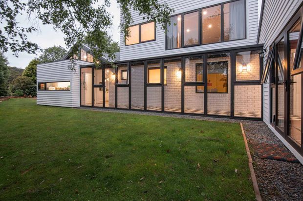 Exterior view of a contemporary multi-level residential extension with white horizontal cladding, dark-framed glazing, and sections of exposed light brick. The building wraps around a lawn and includes extensive full-height windows, warm interior lighting, and a narrow gravel-edged walkway. Mature vegetation surrounds the property.