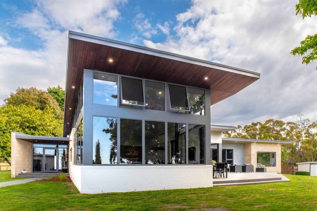 Exterior view of a contemporary single-storey home with a dramatic cantilevered flat roof, extensive floor-to-ceiling glazing, white masonry walls, and a warm timber-lined soffit. The house opens onto a lawn with a covered outdoor entertaining area visible to the side.