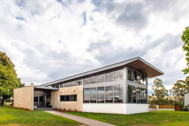 A contemporary two-storey home with extensive floor-to-ceiling glazing, a projecting low-pitched roof with a timber-lined soffit, and pale masonry walls. The building sits in a landscaped suburban setting with a lawn and paved walkway. The glass facade reveals bright interior spaces, while the strong horizontal roofline and exposed structural framing create a modern architectural character.