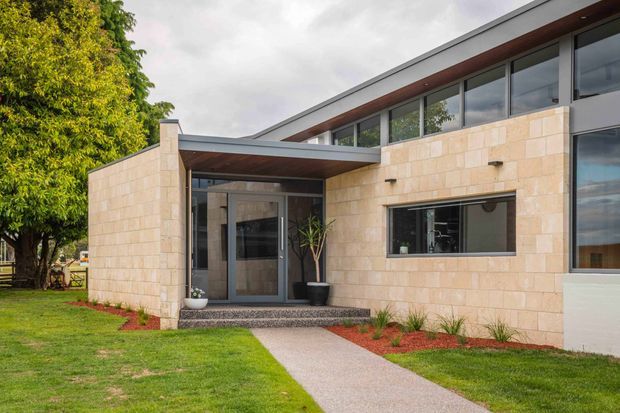 Single-storey contemporary residential facade with pale rectangular stone cladding, extensive dark-framed glazing, and a low-pitched roof with deep timber-lined eaves. A covered entry canopy leads to a glazed front door, approached by a concrete path through a landscaped lawn and garden beds.