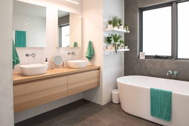 Bright contemporary bathroom featuring a floating timber vanity with two vessel basins, wall-mounted taps, large mirrors, open shelving with plants, and a freestanding white bathtub. The room combines pale timber, white sanitaryware, dark grey tiled walls and floor, black-framed windows, and turquoise towels.