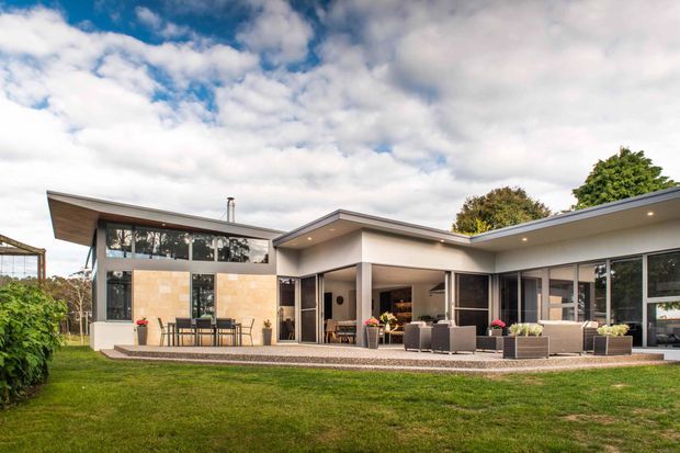 A single-storey contemporary home viewed from the rear garden, with a low-slung asymmetrical roofline, broad eaves, extensive floor-to-ceiling glazing, and an open-plan indoor-outdoor living area. The covered terrace includes outdoor seating and dining, while the lawn and planting frame the building. The facade combines light rendered surfaces, dark window framing, and a warm timber-clad section beneath the projecting roof.